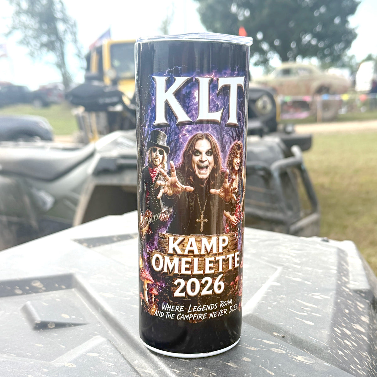 A 20 oz stainless steel tumbler of KLT Kamp Omelette 2026 stands on the edge of a truck bed outdoors, with an ATV and grassy field blurred in the background.